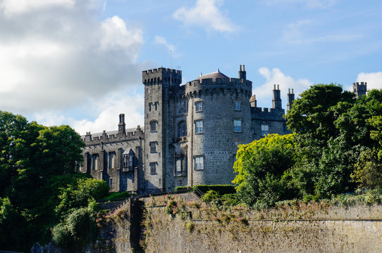 Fortified castle with grey stone walls surrounded by green trees under a blue sky with white clouds. Kilkenny Castle in Ireland.