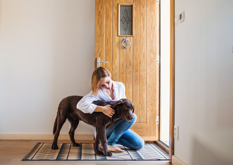 A young woman sitting indoors by the door on the floor at home, playing with a dog.