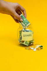 Donation jar with paper money and coins on yellow background. Pills and medicine. Womans hand putting dollar bill into the jar