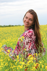 Naklejka na ściany i meble A smiling girl with long hair stands half a turn in a field with crops of blossoming colza in the summer on a sunny day. Close-up.