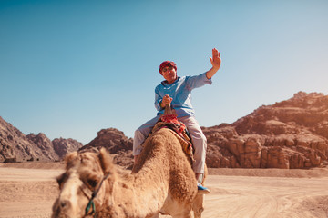Senior man rides a camel in desert by Sinai mountains. Happy tourist waving at camera