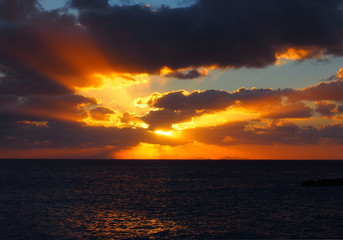Fototapeta na wymiar sunset over a calm dark sea with orange rays shining though dark dramatic evening clouds