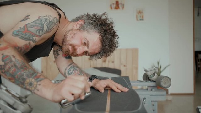 Side view of skateboarder with tattooed arms scrapes side of longboard with brand new grip tape during process of regripping. Recycling and reusing old objects. Handsome millennial hipster at home