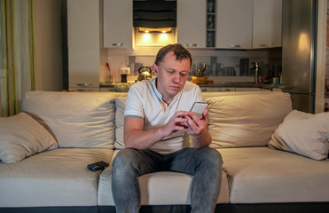 Obraz premium Bored young man sits on the sofa in the evening at home and looks at his phone