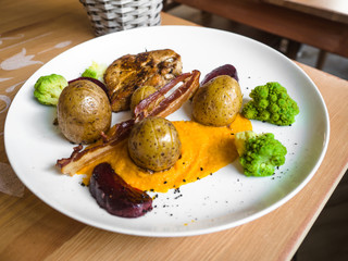 Grilled chicken leg with baked potato and vegetables, carrot cream sauce, bacon, fusion food concept on wooden table in a restaurant.