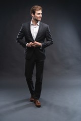 Full length portrait of a handsome young businessman dressed in suit isolated over gray background, posing, looking away, walking