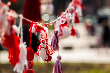 Many of red and white beautiful martisor and martenitsa hanging on the branches of the tree in windy weather during the spring celebration with urecognizable people at the background