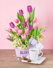 Bouquet of fresh tulips in a wooden container and a cup of black coffee on a pink background.