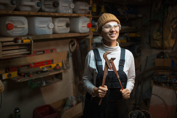 Portrait of smiling young builder girl standing in old workshop room, wearing construction glasses.