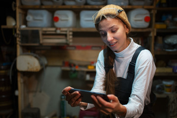 Portrait of young builder girl standing in workshop use smartphone, wearing construction glasses.