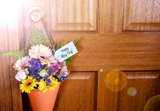 Happy May Day traditional gift of Spring flowers in orange cone hanging from door handle on wooden door, with lens flare.