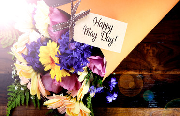 Happy May Day traditional gift of Spring Flowers in orange paper cone on dark wood table with lens flare.