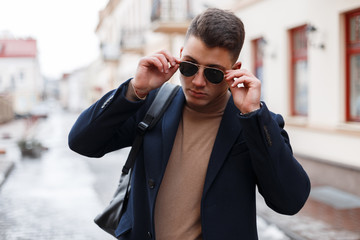 Handsome stylish young hipster man with a stylish hairstyle in a black coat in trendy sunglasses in...