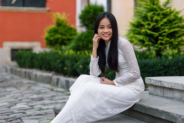 Close up portrait of beautiful asian girl dressed in Ao Dai white dress sitting on the stairs in courtyard.