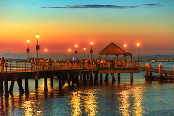 Scenic sunset view of Coronado Ferry Landing on Coronado Island, California, USA. People and tourists walking on old wooden pier reflecting in San Diego Bay at twilight.