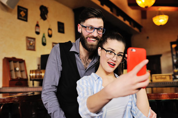 young couple - guy and girl posing and grimacing on the smartphone's camera, making selfie on the background of the bar.