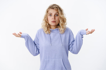 What do I know. Portrait of confused and unaware cute european blond woman with blue eyes standing questioned with clueless gaze shrugging and spread hands sideways in dismay uncertain over white wall
