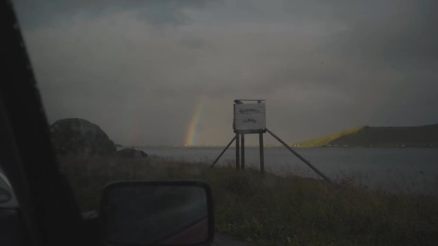 Inside a car view looking out the side window over a stunning lake in the Norwegian countryside with a beautiful rainbow on the horizon on an rainy overcast day in Norway
