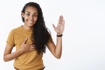 I swear geez. Portrait of cute and friendly happy young nice african-american girl with curly hair raising palm and holding hand on heart as making oath smiling joyfully over gray background