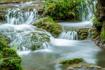 Panoramic view of small waterfalls streaming into small pond in green forest in long exposure 