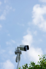 A white security CCTV camera surrounded by trees ready for recording in a bright morning with clear sky.