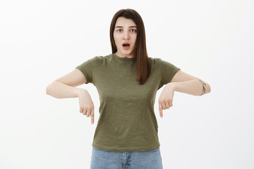 Omg look there. Portrait of concerned and shocked surprised young female open mouth, dropping jaw from amazement and stupor pointing down at stunning impressive product over gray background