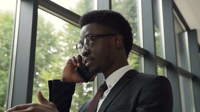 Close up shot of successful afro american businessman in a suit with tie standing near panoramic windows of office building and talking on the mobile phone. Office worker talking the phone. Technology