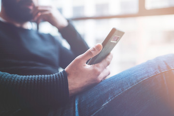 Bearded hispanic man in black pullover using mobile phone application for mobile blog. Confident young hipster guy read news via cellphone from social network at modern loft.Blurred background