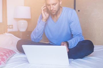 Handsome coworker man working at living room at home. Man sitting at bed using laptop and mobile phone. Blurred background.Flares