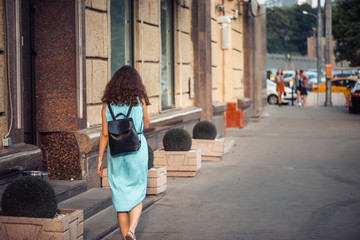 Back view of a hipster girl in a blue dress with black leather backpack walking on city street