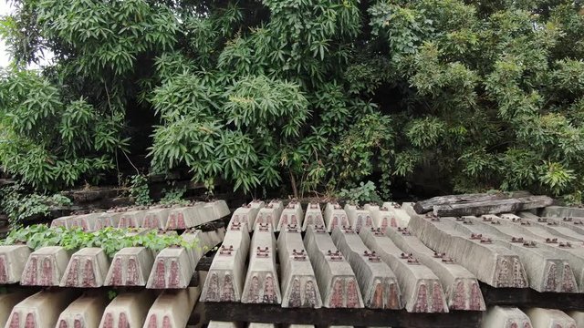 Aerial low view pile of newer concrete and older wooden railroad ties in a railyard in rural tropical Asia; ties are overgrown with foliage. Camera ascends to wide view.