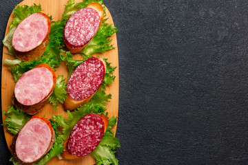 Several sandwiches with sausage and salami and sauce on a black board, flat lay background with copyspace