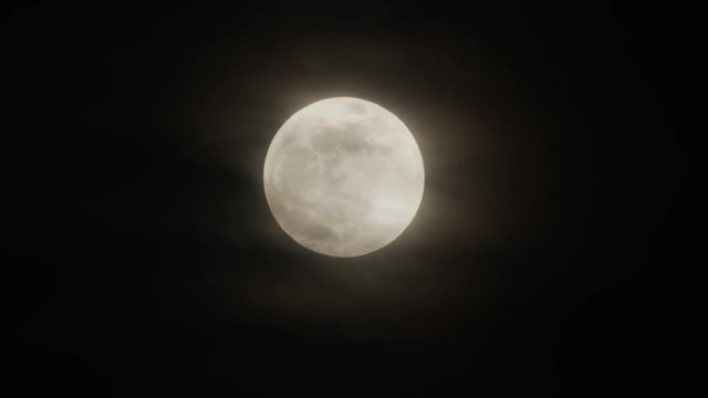 Wispy clouds pass from left to right in front of a bright rising full super moon.