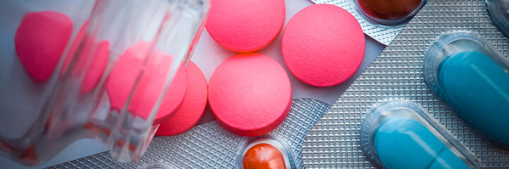 Pink pills are poured from a glass jar and blisters with colored tablets are scattered on a white background. The view from the top.