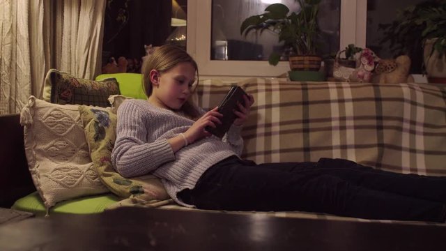 The girl is lying on the sofa and reading an e-book in