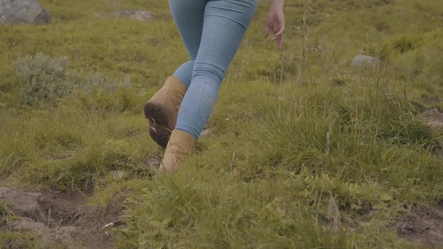 Woman's feet and shoes hiking up a grassy mountain with boulders and green vegetation on an overcast day in Norway