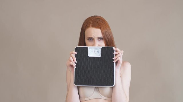 Anorexic girl with a sad face holds the scales and looks at camera, on a gray background.