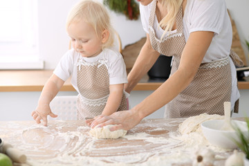 Little girl and her blonde mom in beige aprons  playing and laughing while kneading the dough in kitchen. Homemade pastry for bread, pizza or bake cookies. Family fun and cooking concept