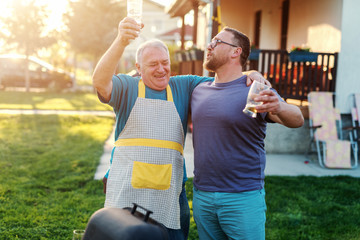 Cheerful father in law and son in law hugging and drinking beer while standing next to grill in...