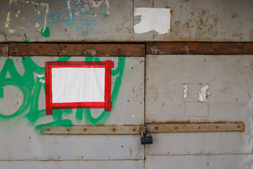 A sheet of white paper glued with red adhesive tape to a locked metal gate. On a dirty iron wall are scraps of ads and paint stains. Perfect for background and grunge design. Place for text.