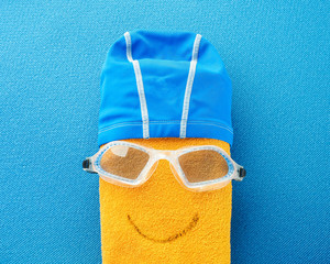 Naklejka premium Flat lay composition with Goggles and swimming cap, yellow towel in the form of a smiling positive face. equipment for swimming pool on blue background. top view. Swimming set.