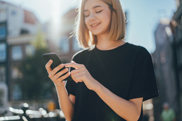 Happy smiling hipster blonde girl browsing news feed of her mobile phone while walking on the city...