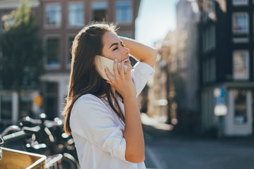 Happy laughing young girl having a conversation talk via mobile cell telephone while walking on the street, talking to her friends and smiling, flare light and city streets on the background