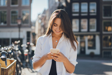 Happy smiling girl walking around the streets using her mobile phone typing to her friend via messenger app and browsing internet, blurred city streets on the background, horizontal