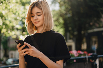 Young pretty hipster girl in a black t-shirt using modern smartphone in city for texting friends and browsing internet, smiling and enjoying wireless internet connection, space for your logo or design