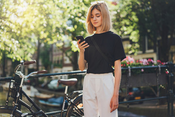 Happy smiling young hipster girl browsing news feed of her mobile phone while walking on the city streets and enjoying wireless connection talking with friends and using internet