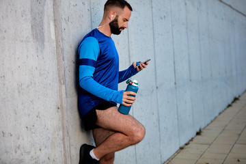 Male runner leaning on wall and using smart phone. In other hand bottle with water.