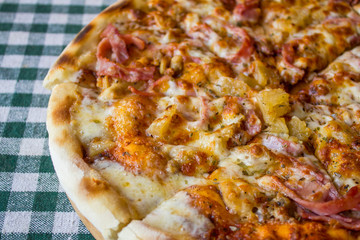 cooked round pizza with bacon and cheese on a checkered tablecloth close up sliced into pieces