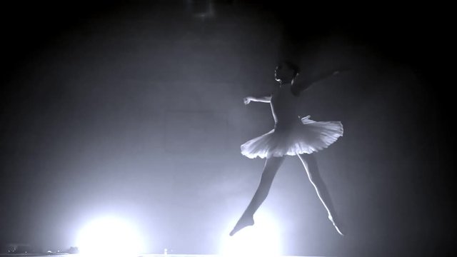 Young ballerina girl in tutu bounces in slow motion moving her legs and crouching at the end in a smoky room with a spotlight in the background
