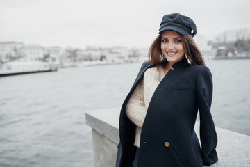 A young beautiful woman with long,hair and gray eyes,beautiful makeup,pink plump lips,sweet smile,brunette,spending her time outdoors in the spring in the city.Portrait of happy young woman in a cap